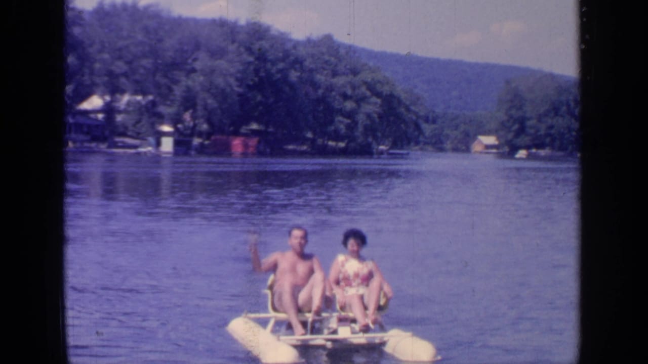 Man and woman ride a yellow paddleboat on a lake as the man waves toward the camera.
