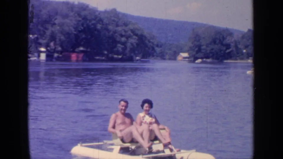 The 1964 Greenwood Lake footage shows the couple relaxed on the paddleboat, framed by blue water, trees, and distant shore structures.