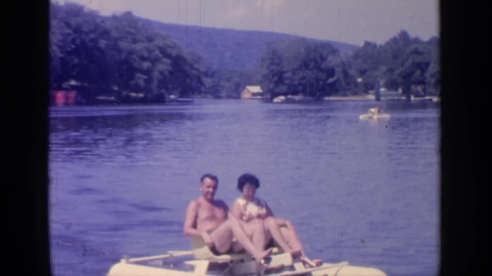 A couple rides a yellow paddleboat on Greenwood Lake, with a distant small boat, trees, shoreline houses, and hills behind them.
