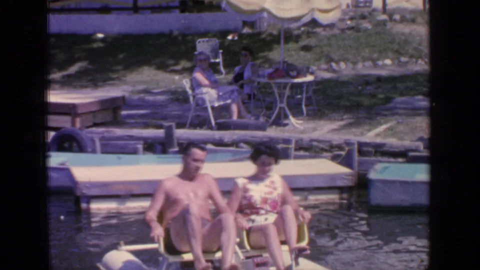 Two adults ride a paddle boat near a dock at Greenwood Lake, with other adults seated under a yellow umbrella on shore.