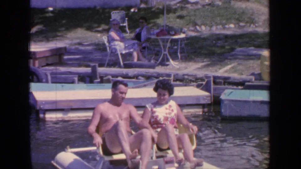 A shirtless man and a woman in a floral dress sit in a paddle boat near the Greenwood Lake dock in 1964 footage.