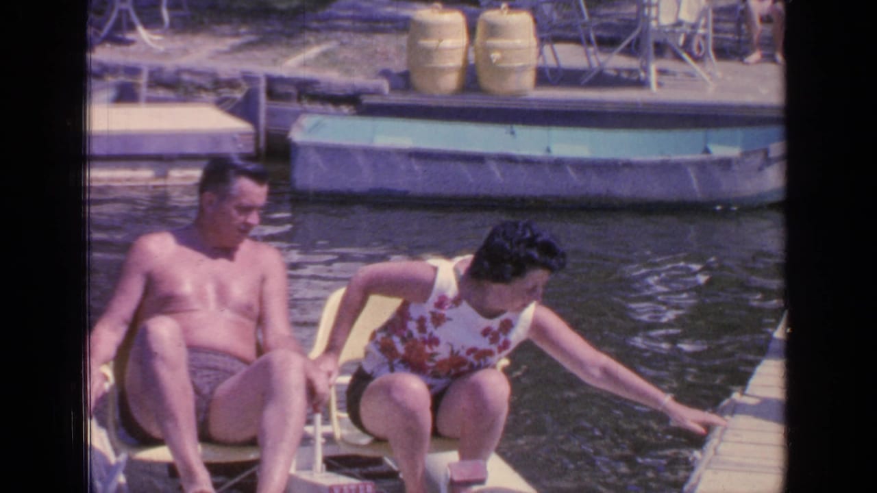 At Greenwood Lake in 1964, the woman reaches from the yellow pedal boat toward the wooden dock while the man sits beside her.