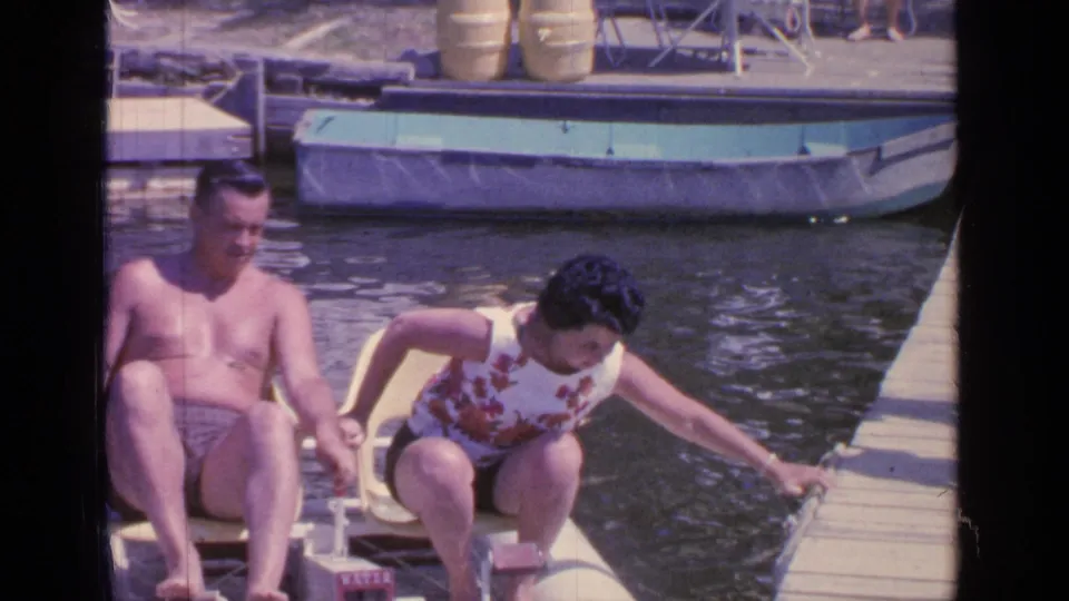 1964 lakeside home-movie frame of a couple in a yellow pedal boat as the woman steadies herself near the dock.