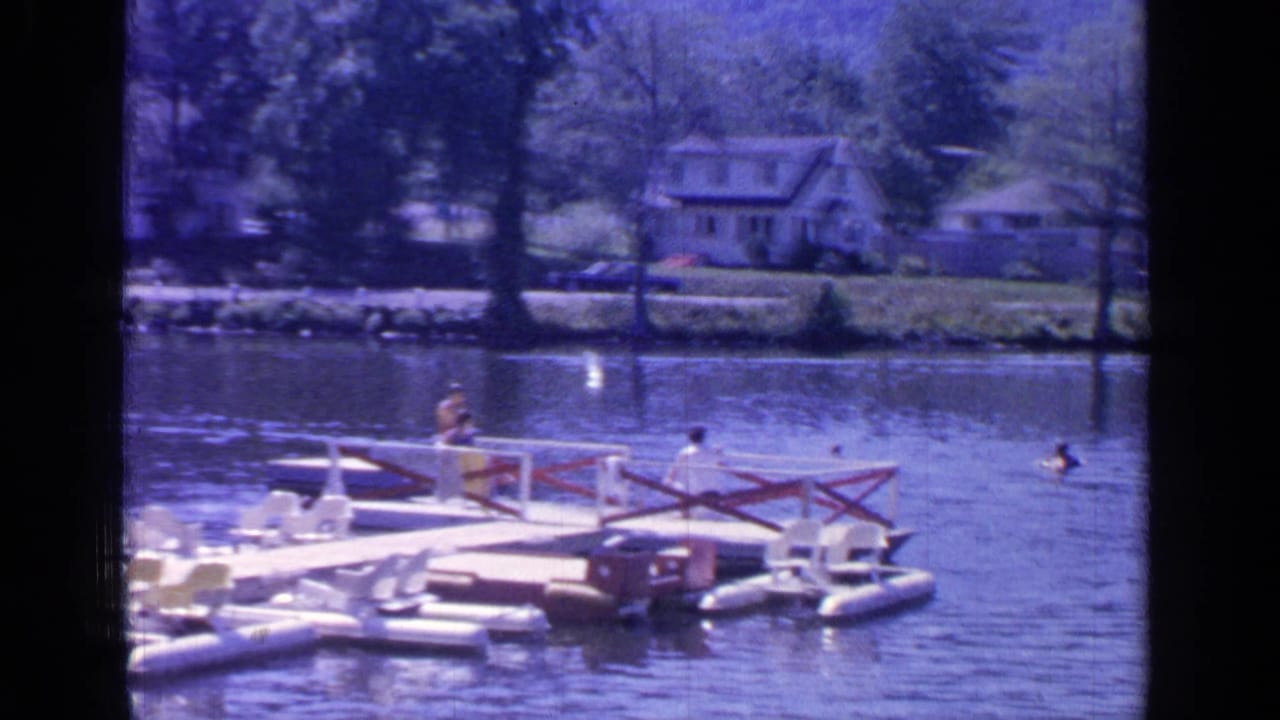 A floating platform sits near swimmers on Greenwood Lake, with trees and shoreline houses across the water.