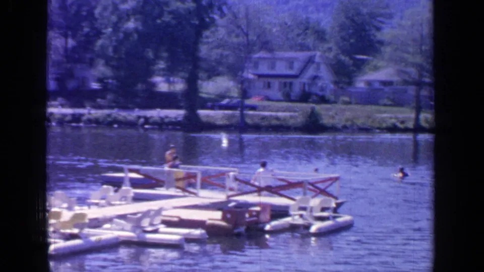 A floating platform sits near swimmers on Greenwood Lake, with trees and shoreline houses across the water.