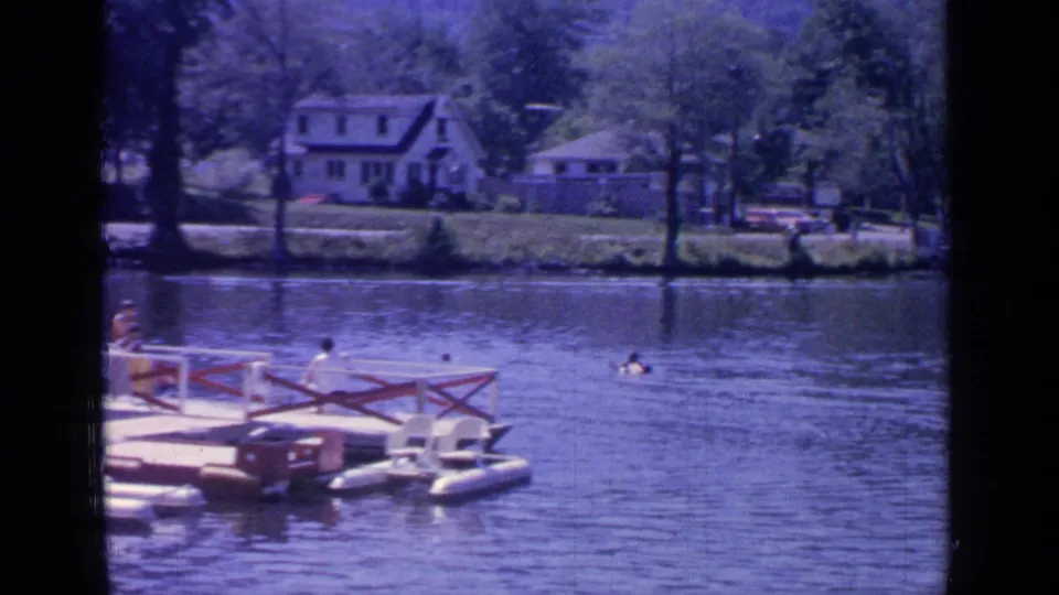 The camera frames swimmers near a small floating platform, with Greenwood Lake residences and trees in the background.