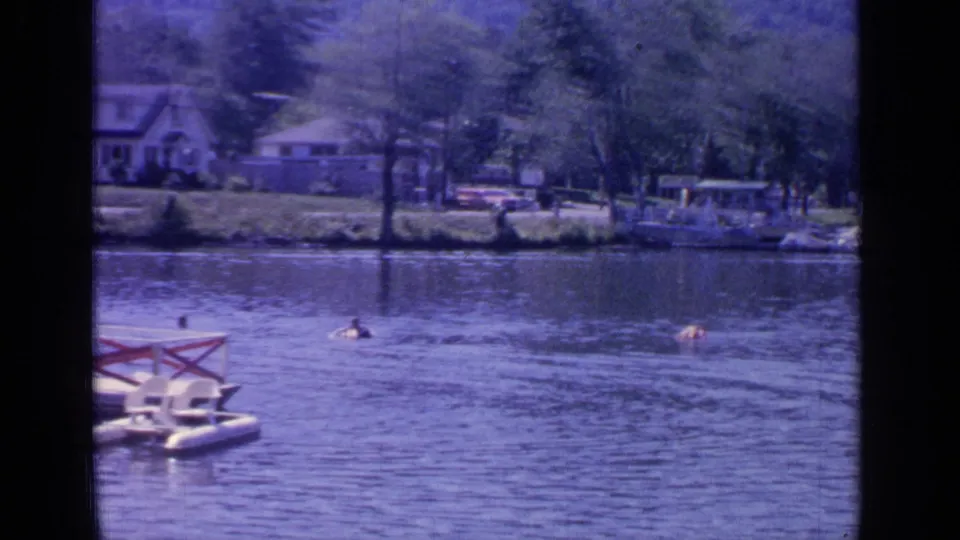 Swimmers cross the water near a small floating platform on Greenwood Lake, with houses and trees beyond the shore.