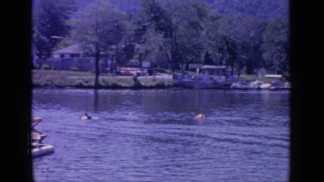 1964 8mm view of Greenwood Lake with swimmers in open water and a wooded shoreline with docks and lakeside houses.