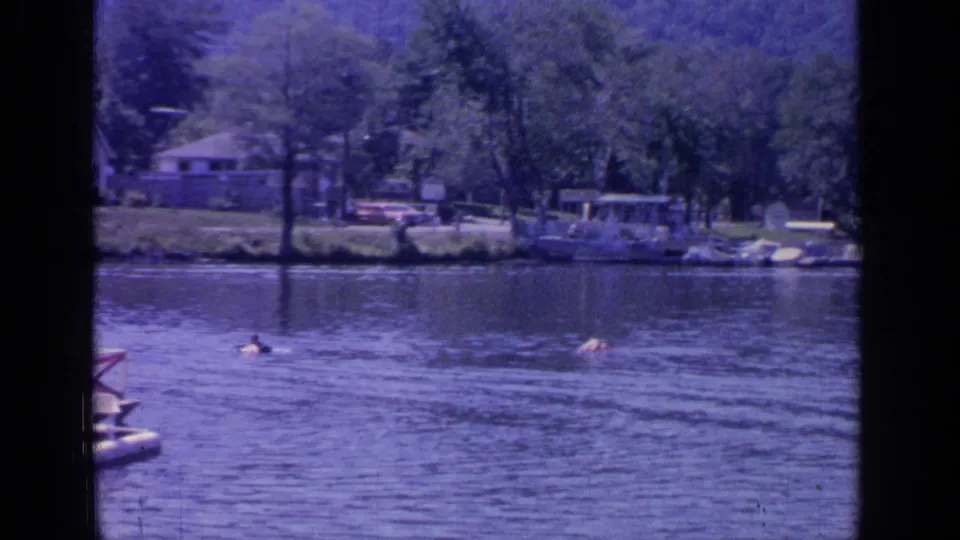 1964 8mm view of Greenwood Lake with swimmers in open water and a wooded shoreline with docks and lakeside houses.
