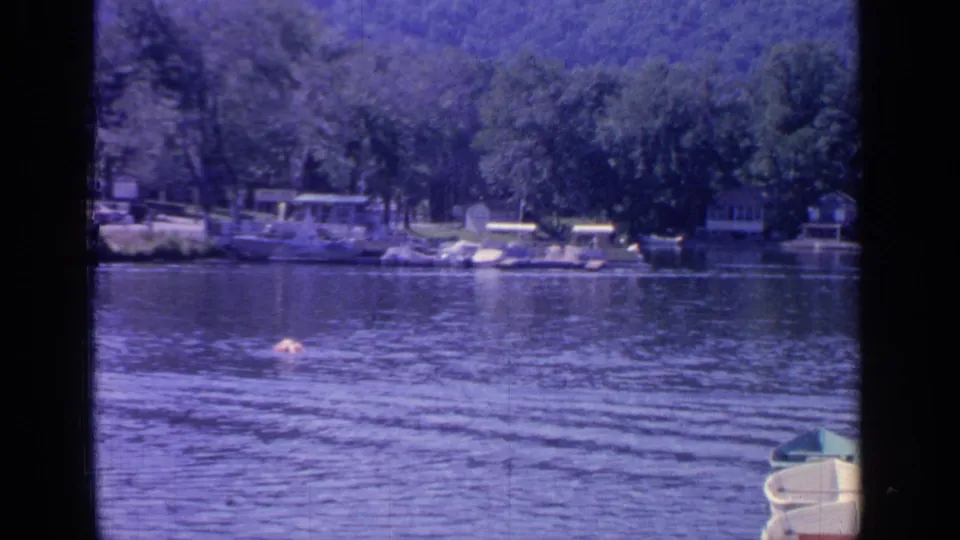 1964 Greenwood Lake home-movie frame showing rippled water, a distant swimmer, boats near shore, and wooded hills.