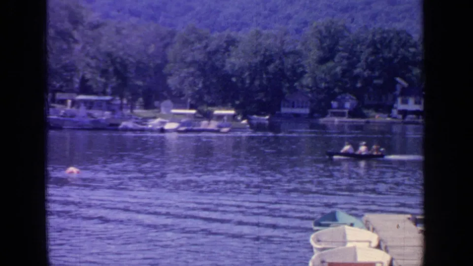 Two people move across Greenwood Lake in a small boat, with docks, moored boats, trees, and houses along the shore.