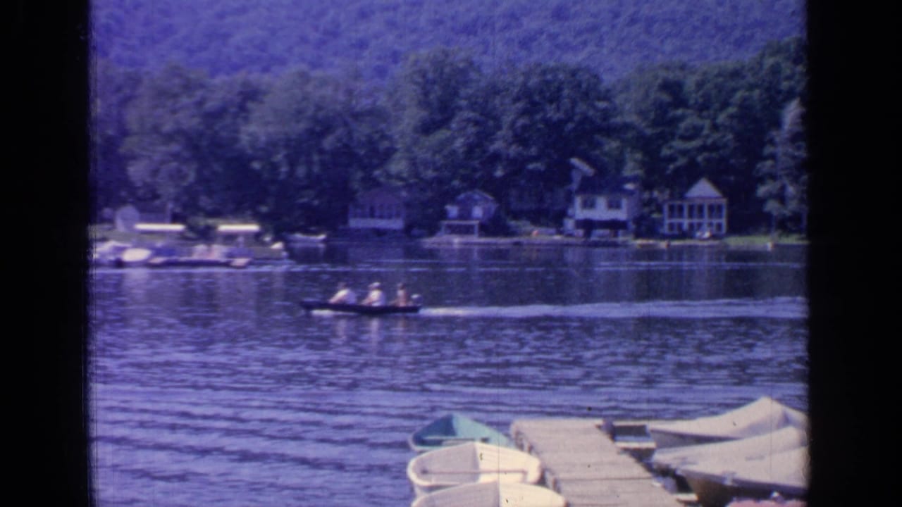 1964 Greenwood Lake footage frame with a motorboat and distant passengers crossing calm water before cottages and wooded shoreline.