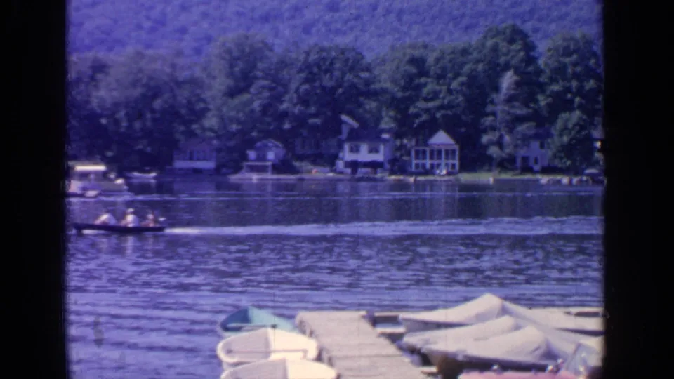 1964 Greenwood Lake frame showing the motorboat partly at left, its wake, shoreline cottages, and small boats tied near a dock.