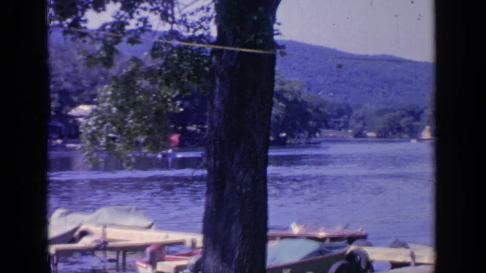 Tree-framed 1964 Greenwood Lake scene with a dock, covered boats, rippled water, and a distant shoreline below wooded hills.