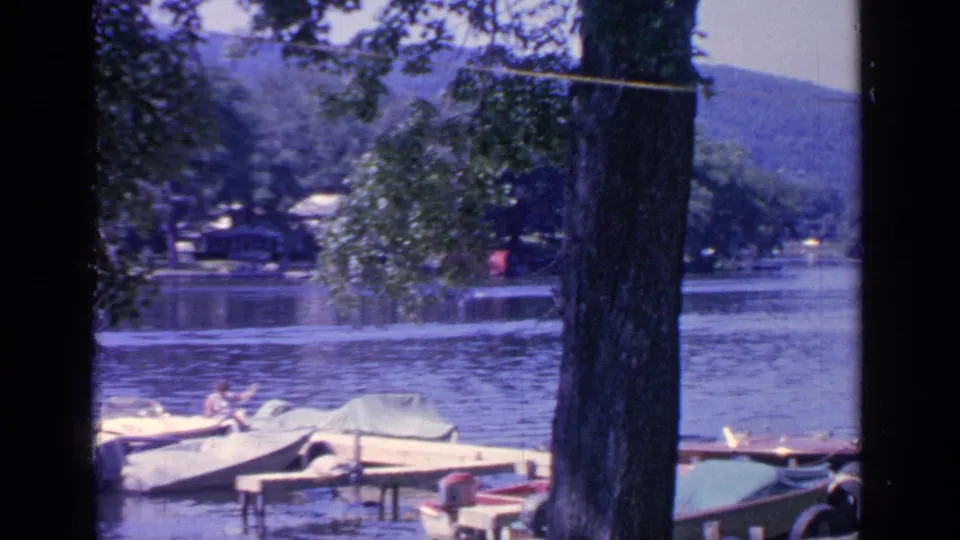 1964 Greenwood Lake dock scene with covered boats, a large tree trunk, calm water, hills, and a distant person in a red shirt.