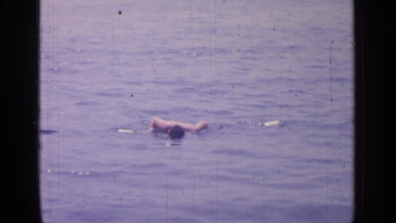 1964 8mm footage at Greenwood Lake shows a person swimming across calm lake water, framed by the dark edges of the film scan.