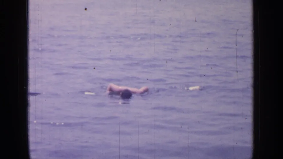 1964 8mm footage at Greenwood Lake shows a person swimming across calm lake water, framed by the dark edges of the film scan.