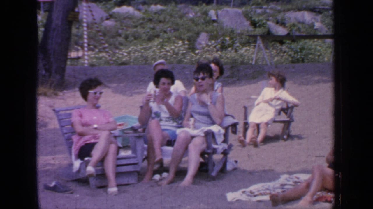 Women sit on wooden lounge chairs on a lakeside shore while another person reclines nearby.