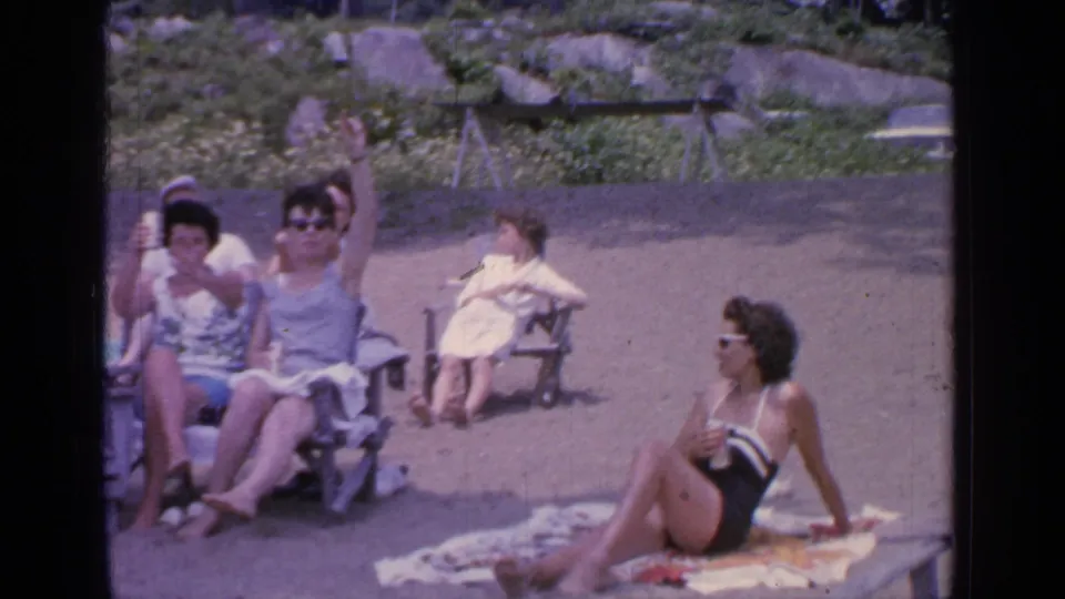 The 1964 Greenwood Lake home movie shows women on the shore, including one waving toward the camera and another sitting on a towel in a dark swimsuit.
