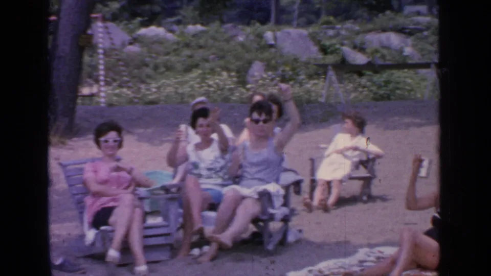 Women relax on wooden chairs at Greenwood Lake in 1964 as one waves toward the camera, with towels, shore, trees, and rocks visible around them.