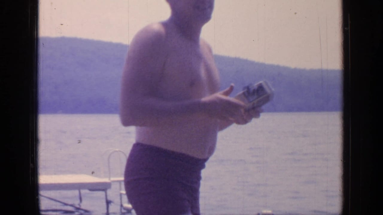 Shirtless man in dark swim trunks standing near a lake with a dock ladder behind him.