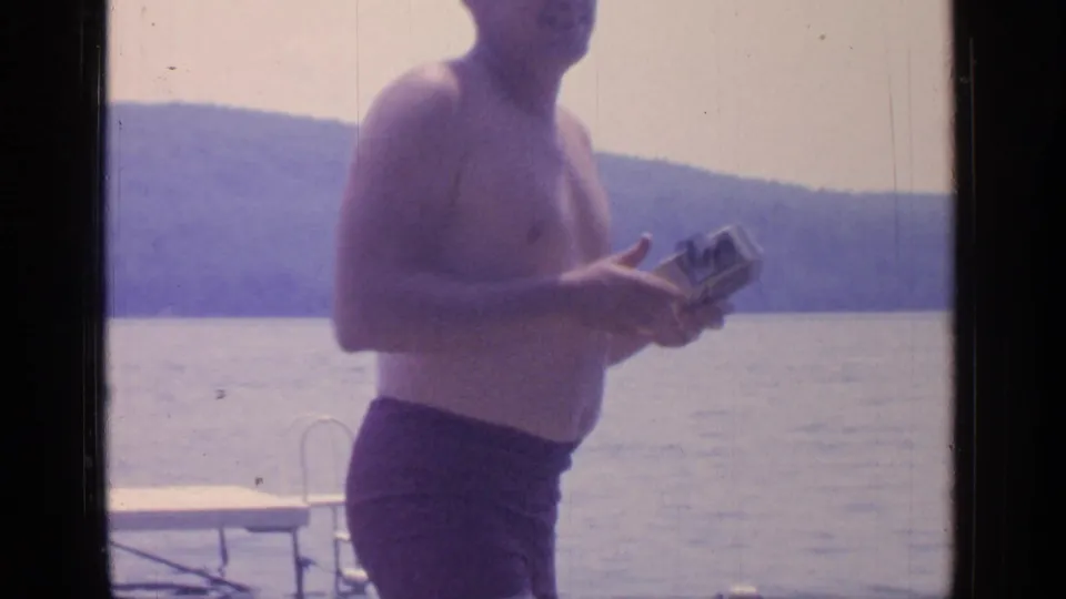 At Greenwood Lake in 1964, a shirtless man in dark swim trunks stands by the water holding a small object.