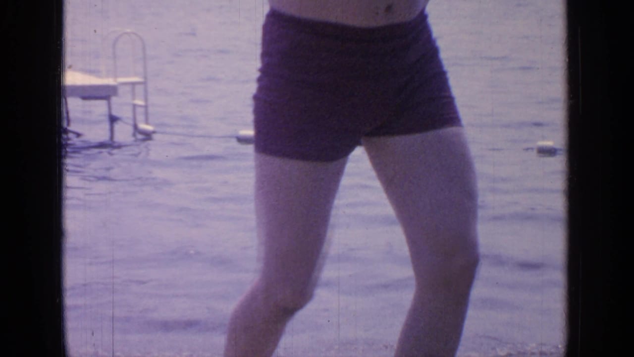 A man in dark swim trunks stands close to the lake, with rippling water and a small dock ladder in the background.