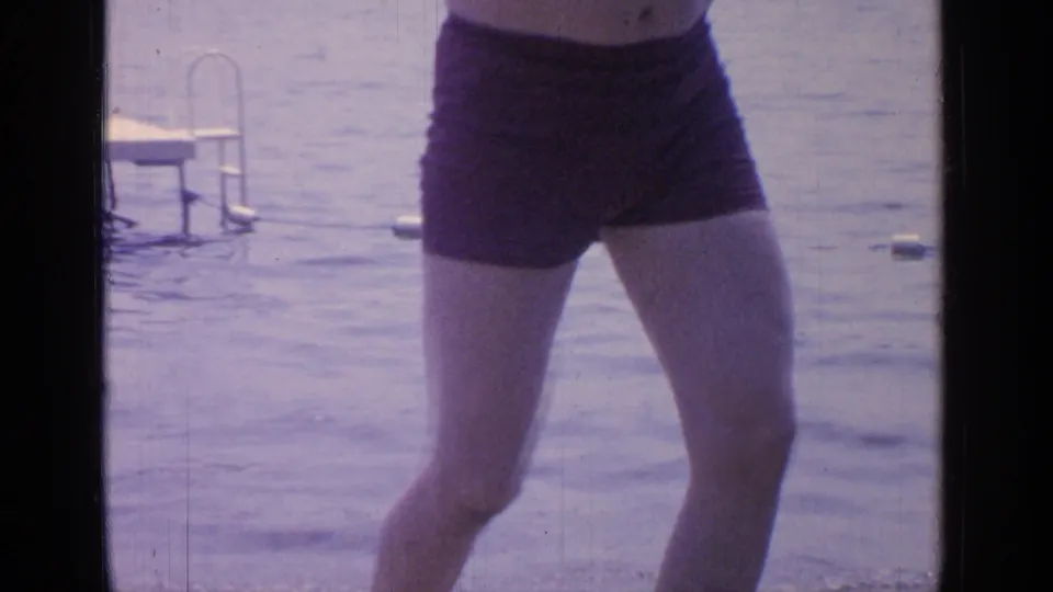 A man in dark swim trunks stands close to the lake, with rippling water and a small dock ladder in the background.
