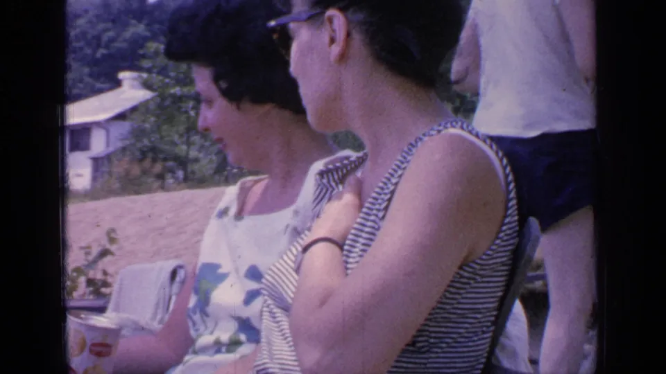 Two women relax in lawn chairs at Greenwood Lake in 1964, one holding a can while a man stands partly behind them.