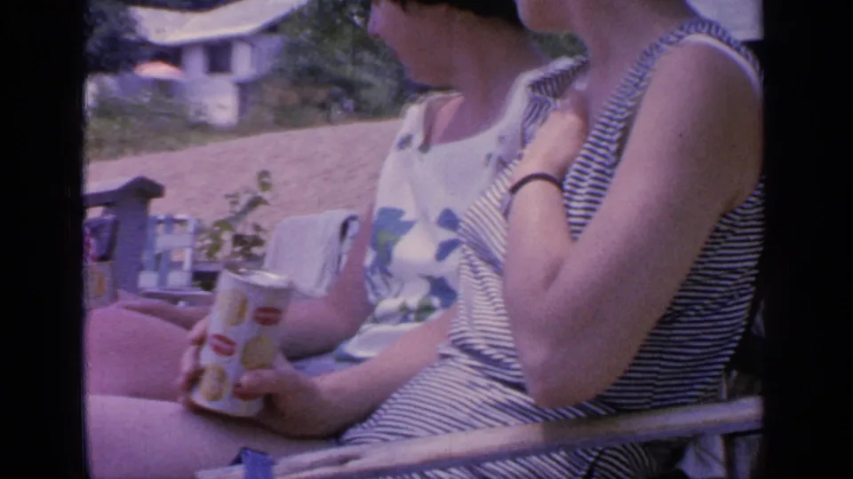 A woman in a striped sleeveless top sits in a lawn chair at Greenwood Lake in 1964, holding a branded drink can.