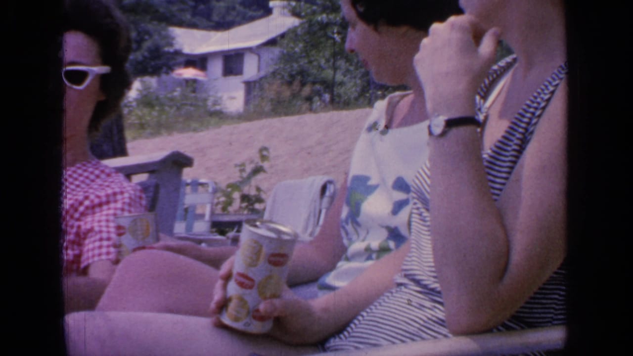 Three women sit outdoors by a lake, with sunglasses, summer clothing, soda cans, and a hillside cottage behind them.