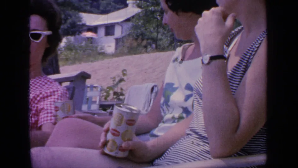 1964 Greenwood Lake home-movie frame showing three women relaxing on wooden seating with soda cans, summer clothes, and a cottage on the slope behind them.