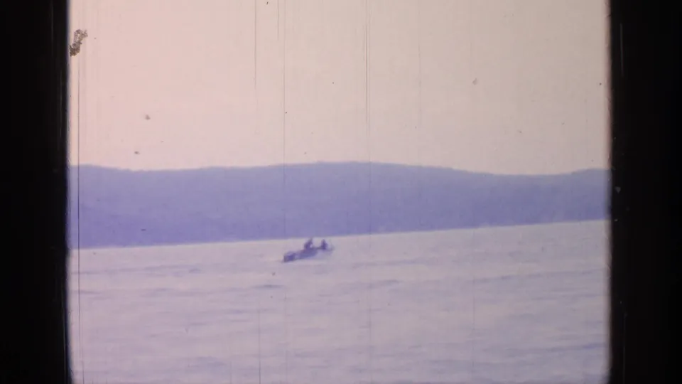 1964 Greenwood Lake frame showing a small boat on open water beneath distant hills, rendered in faded 8mm color.