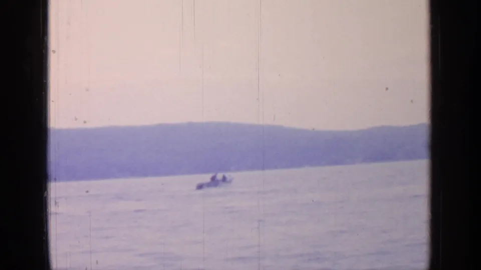 1964 Greenwood Lake 8mm frame showing a small motorboat on calm water, distant wooded hills, pale sky, and dark window edges.