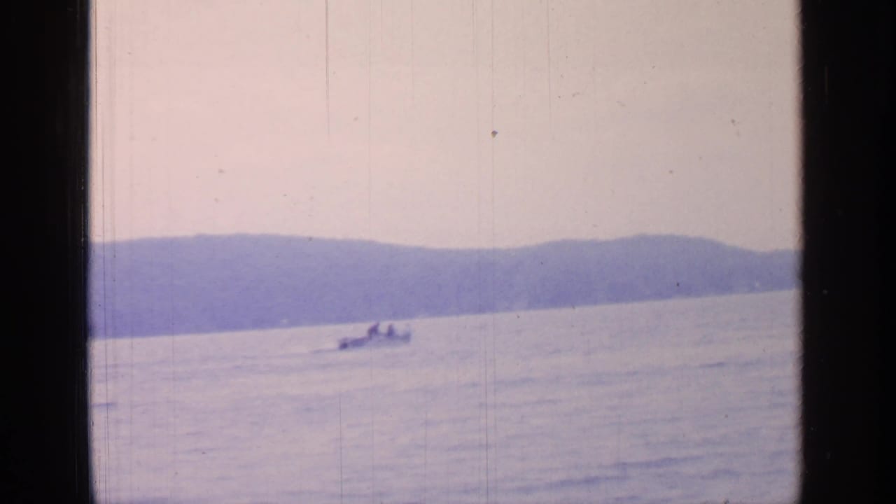 1964 Greenwood Lake frame with a small boat and distant people, soft horizon hills, pale sky, and visible 8mm film wear.