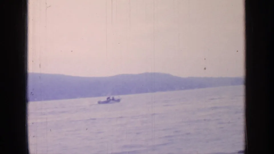 Greenwood Lake home-movie frame from 1964 showing a small motorboat, distant figures, calm water, hills, and scratched 8mm texture.