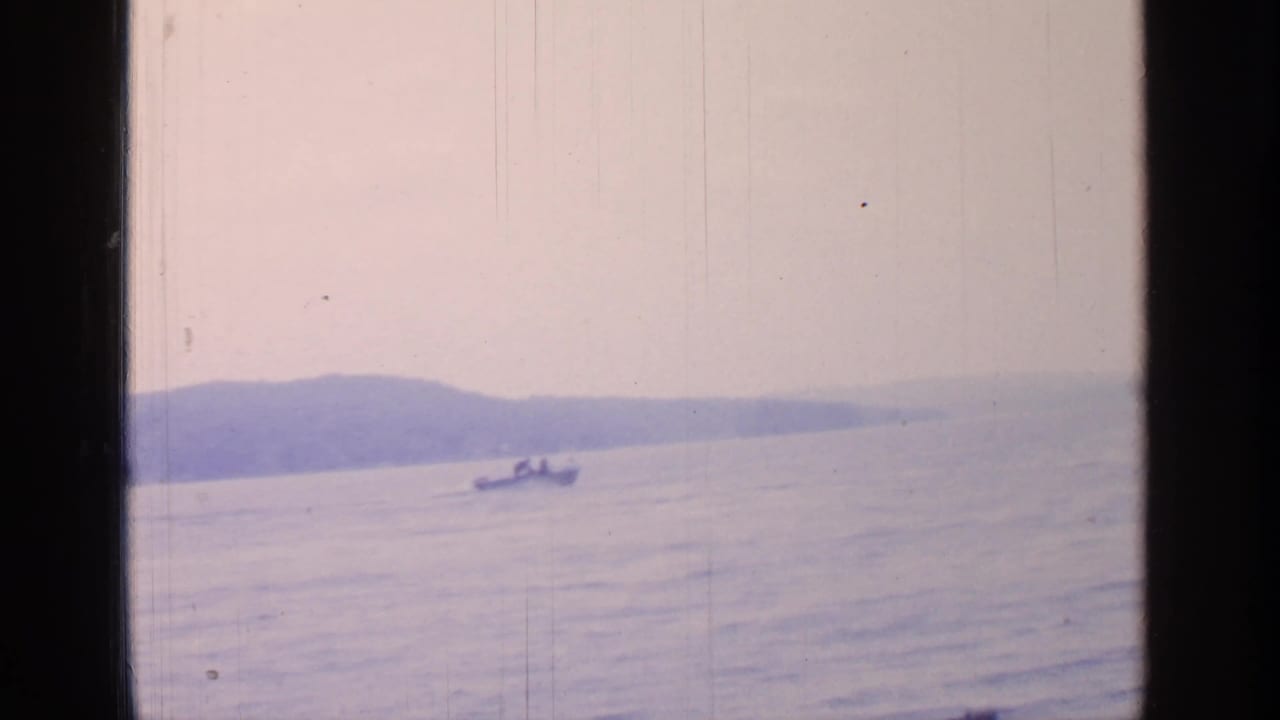 1964 Greenwood Lake view of a small boat crossing calm water, framed by a dark window edge and distant wooded shoreline.