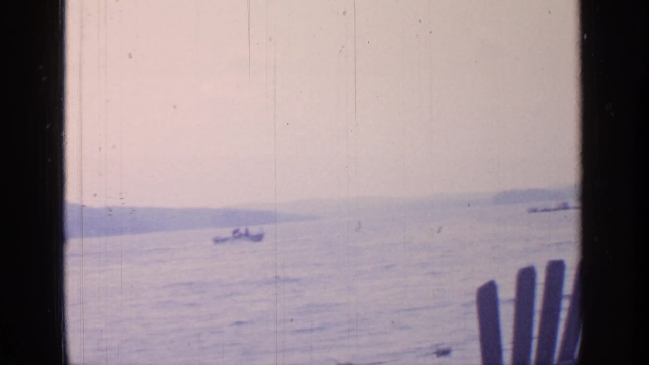 Boat on open lake water with dark window edges and part of a wooden chair at the right.