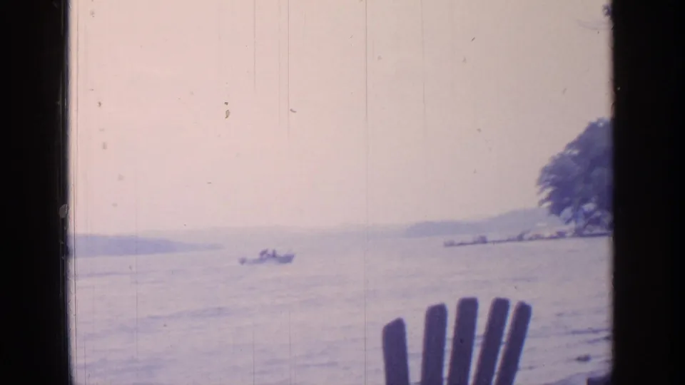 Greenwood Lake home-movie frame showing a small boat, tree-lined shore, cottage, and foreground wooden chair in 1964.