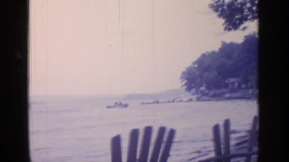 1964 Greenwood Lake scene with a small boat on the water, shoreline trees, cottages, and wooden chairs in the foreground.