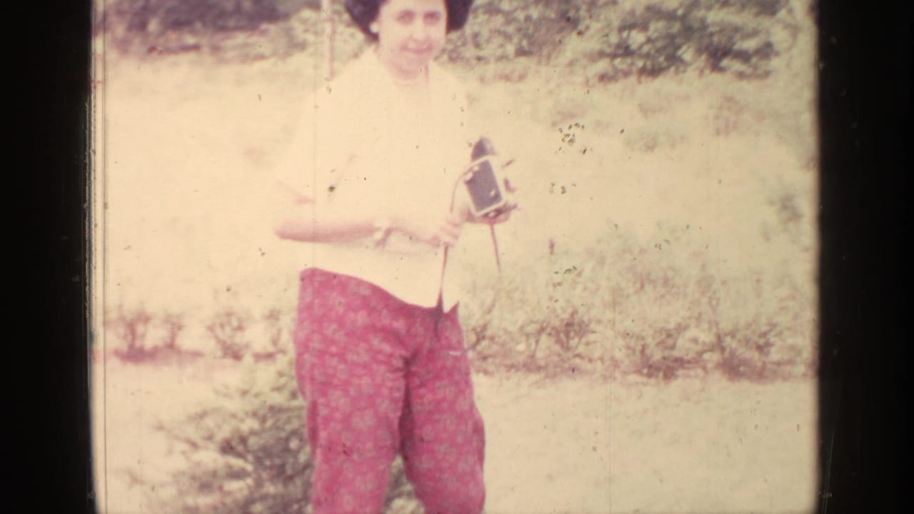 Woman outdoors holding a small film camera beside grass and shrubs.