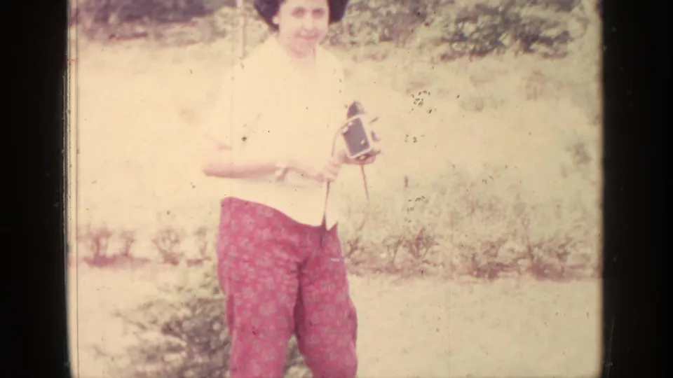 Adult woman in Greenwood Lake home-movie footage, 1964, standing outdoors with a film camera and red patterned pants.
