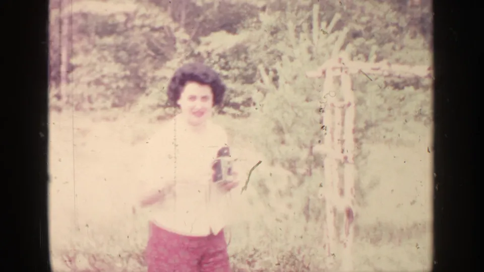 Adult woman smiles toward the lens in 1964 Greenwood Lake footage while holding a small film camera outdoors.