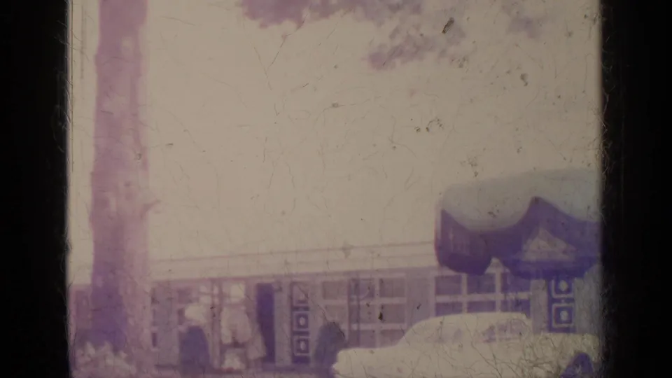Two adults appear outdoors near trees and a low building, likely in Greenwood Lake, New York, in 1964 home-movie footage.