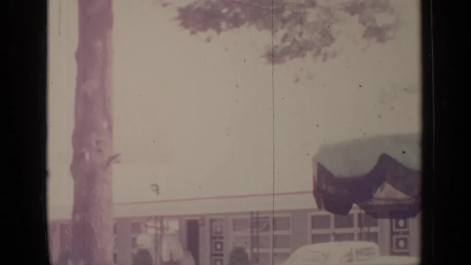 A low mid-century building appears behind trees, with a dark awning or roof detail in faded 1964 home-movie footage.