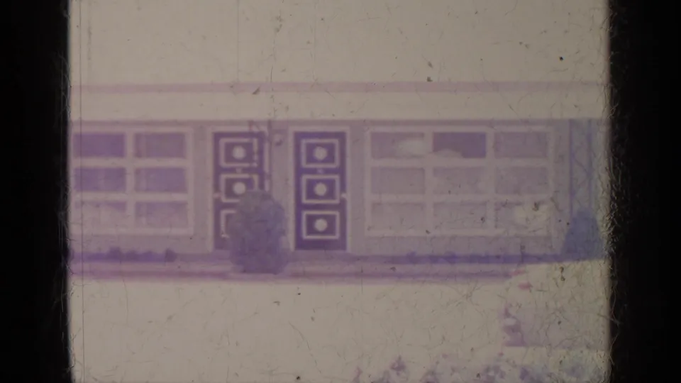 The building facade shows large windows, square-patterned doors, and a garage-like opening in faded Greenwood Lake home-movie footage.