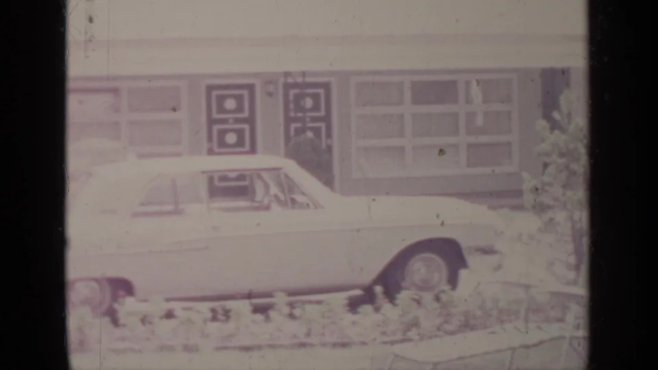A light-colored classic American car is parked outside a low building with patterned doors and windows, likely Greenwood Lake in 1964.