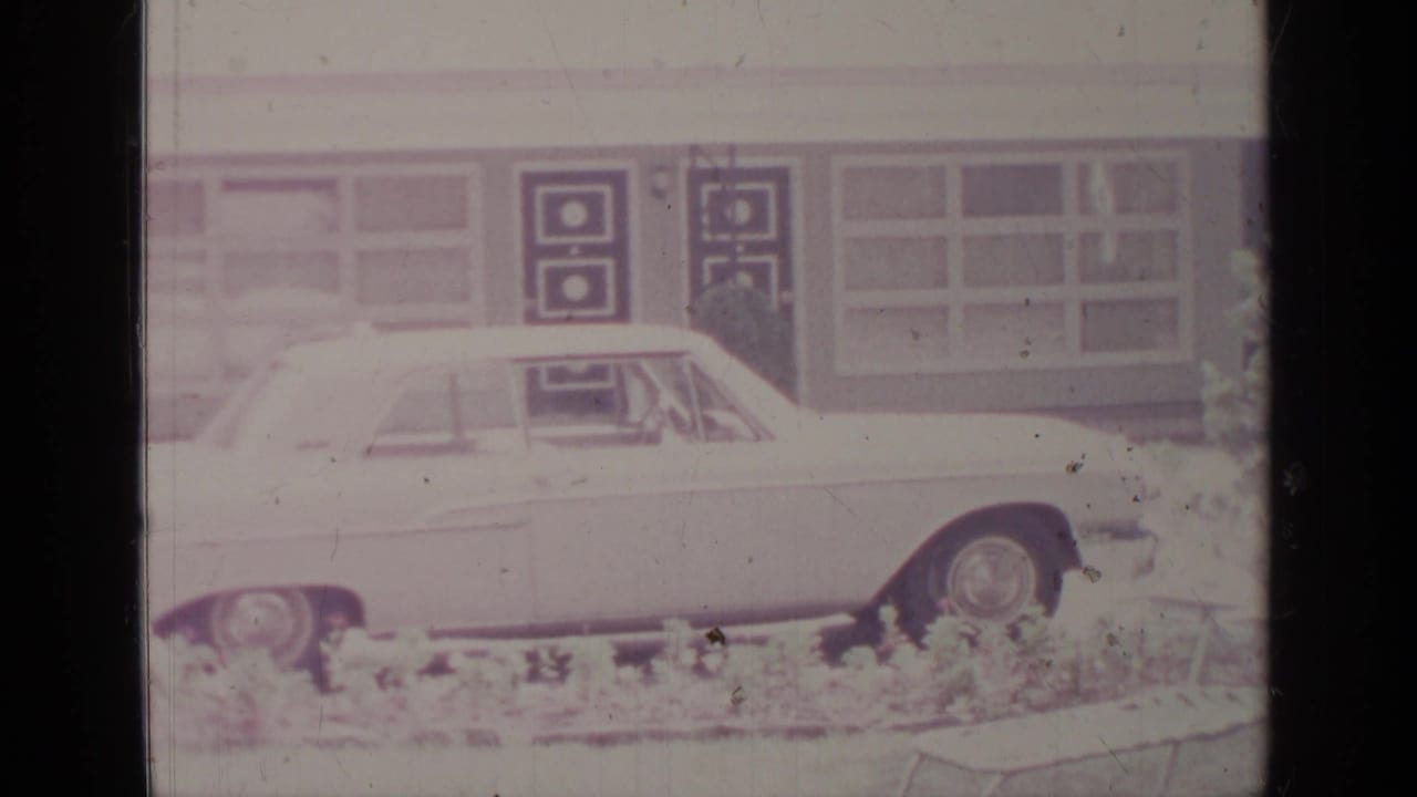 1964 Greenwood Lake 8mm frame showing a white sedan beside a low mid-century building facade with windows and geometric door panels.