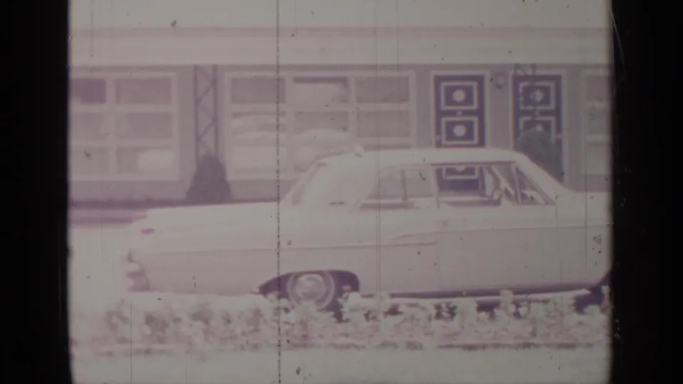 Final 1964 frame showing the rear side of the white sedan near a building with rectangular windows and dark patterned doors.