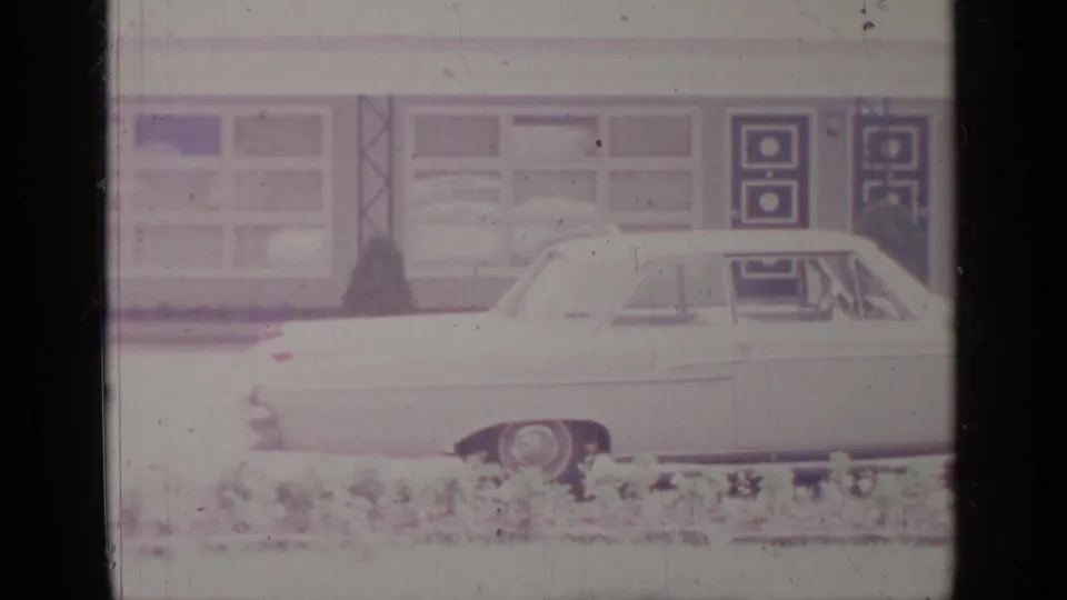 1964 Greenwood Lake 8mm frame showing a light-colored sedan beside a low building with large windows and geometric door panels.